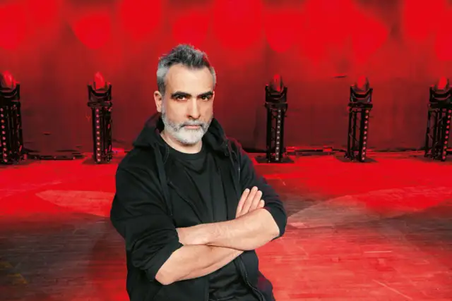 Abelard Giza stand up, pewny siebie, z rękami skrzyżowanymi na piersi, na tle czerwonych świateł scenicznych.