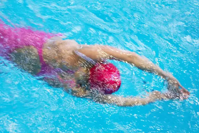 Pływanie kraulem to świetny trening całego ciała, poprawia kondycję i samopoczucie. Widoczna osoba w różowym stroju i czerwnym czepku pokonuje dystans.