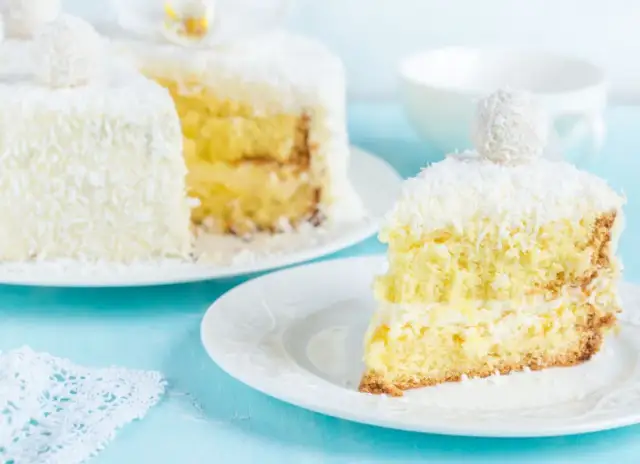 Pyszne ciasto rafaello na biszkopcie, obsypane wiórkami kokosowymi. Kawałek na talerzyku kusi kremowym nadzieniem.