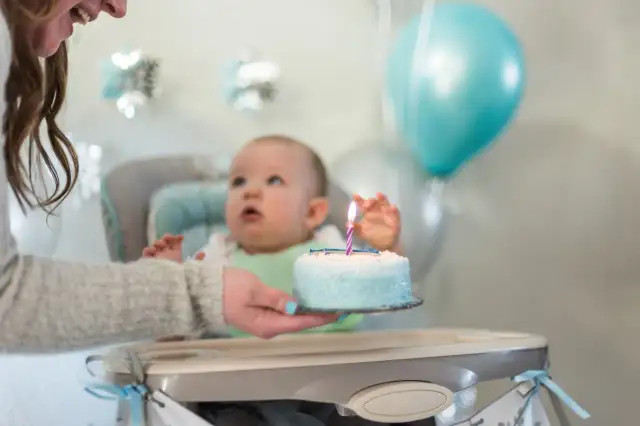 Dziecko w krzesełku z tortem urodzinowym i zapaloną świeczką, balony w tle.