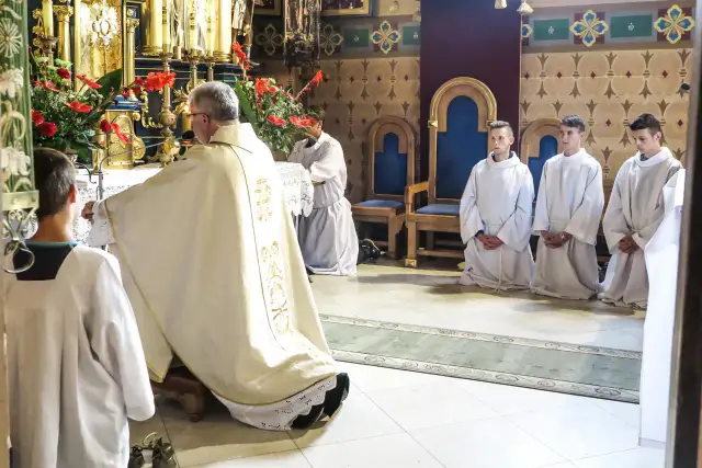Ksiądz w szatach odprawia nabożeństwo, klęczą ministranci w białych albach.