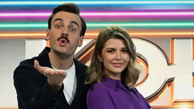 Agnieszka Dziekan, prowadząca Koło Fortuny, uśmiecha się do kamery. Obok niej stoi mężczyzna z wąsami.