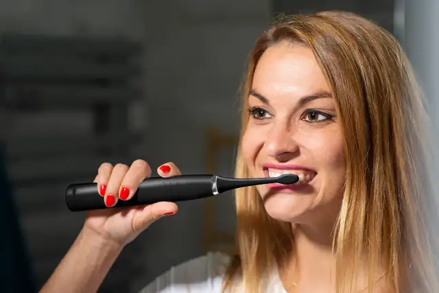 Kobieta z czerwonymi paznokciami używa czarnej szczoteczki sonicznej do zębów.