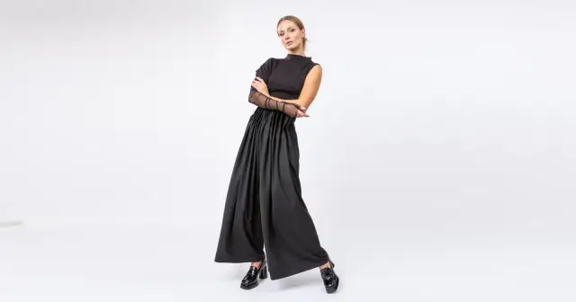 Kobieta w czarnym, eleganckim stroju z szerokimi spodniami i siateczkowymi rękawami, pozuje na białym tle.