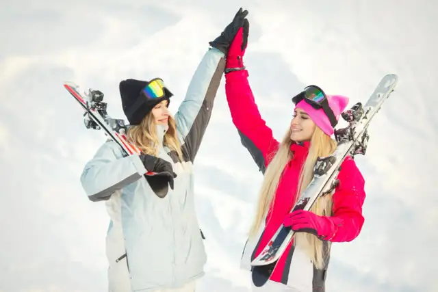 Dwie dziewczyny w zimowych kurtkach i goglach cieszą się na stoku, trzymając narty damskie.