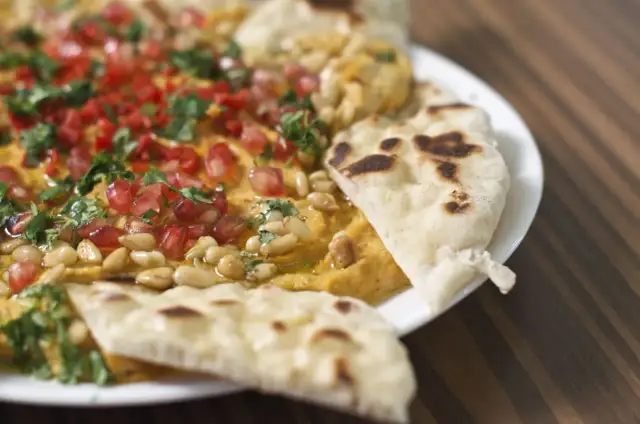 Chlebek naan z hummusem, granatem, orzeszkami pinii i pietruszką na białym talerzu.