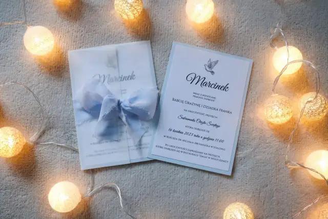 Dwa zaproszenia na chrzest, jedno w kopercie z błękitną wstążką, otoczone girlandą świetlną.