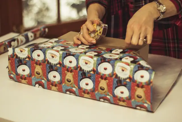 Ręce owijają prezent świątecznym papierem z Mikołajami i bałwankami, używając taśmy klejącej.