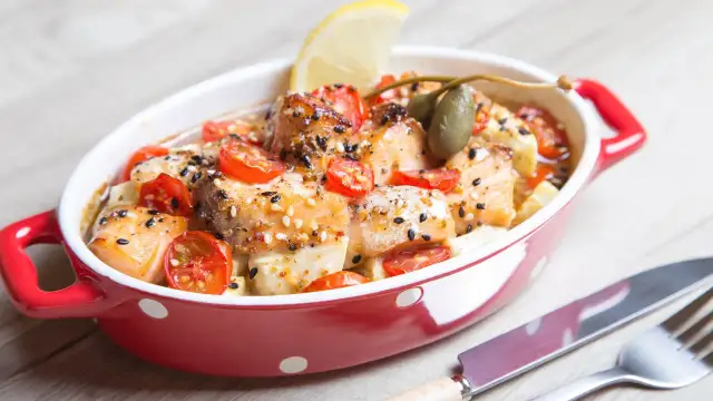 Pieczony łosoś z pomidorkami koktajlowymi, kaparami i cytryną w czerwonym naczyniu.