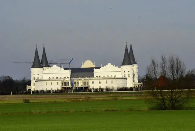 Biały budynek przypominający zamek z ciemnymi wieżami, w tle żurawie budowlane.