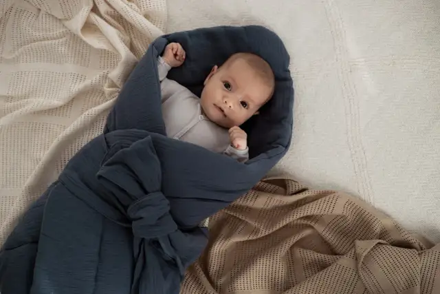 Niemowlę wtulone w granatowy rożek, otoczone jasnymi i beżowymi kocykami.