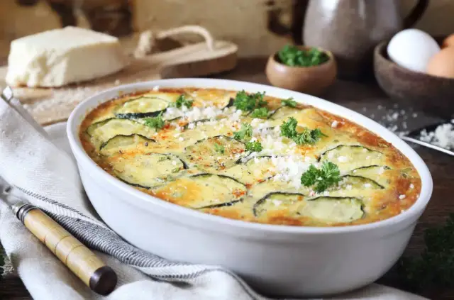 Zapiekanka z cukinii w białym naczyniu, ozdobiona natką pietruszki i parmezanem.