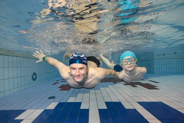 Dwóch pływaków pod wodą, jeden z nich uczy drugiego, jaki styl pływania jest najzdrowszy.