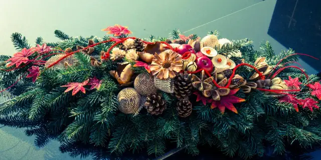 Podłużny stroik cmentarny z gałązek iglastych, szyszek, suszonych kwiatów, jabłek i czerwonych liści.