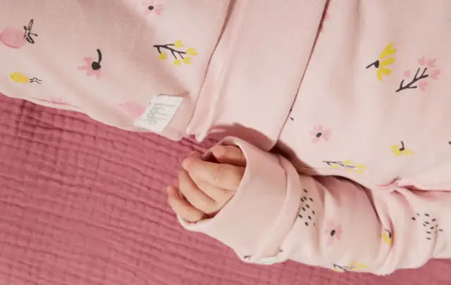 Dłoń niemowlaka w różowym pajacyku w kwiatki, na tle różowej tkaniny.