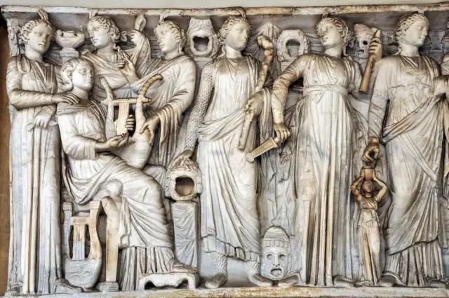 Kamienna płaskorzeźba przedstawia grupę kobiet, w tym Demeter i Korę, otoczone instrumentami muzycznymi i symbolicznymi przedmiotami.