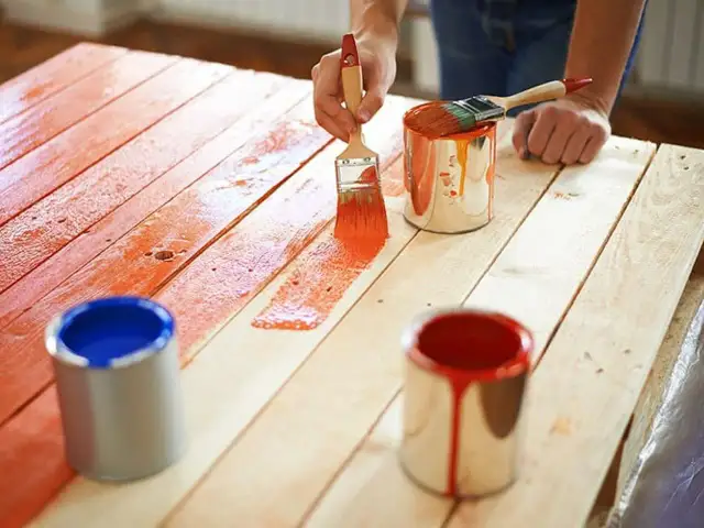 Malowanie na drewnie farbami akrylowymi. Ręce malują deski na pomarańczowo, obok stoją puszki z farbą.