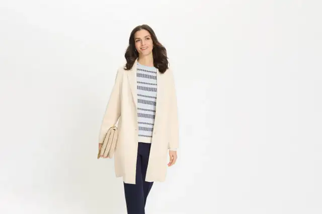 Kobieta w jasnym płaszczu i granatowych spodniach, trzymająca beżową torebkę. Stylowa stylizacja na co dzień.