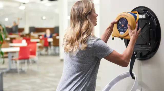 Kobieta zdejmuje żółty defibrylator z uchwytu na ścianie w jasnym pomieszczeniu.