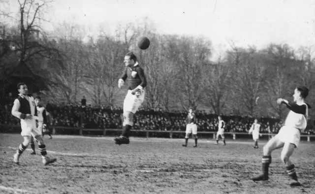 Piłkarz wyskakuje do główki, by trafić w piłkę. Na boisku widać innych graczy i trybuny pełne widzów.