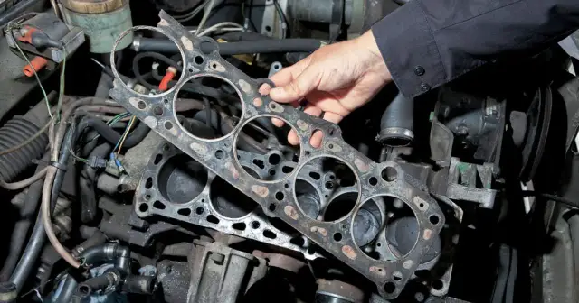 Uszkodzona uszczelka pod głowicą, widoczna na tle silnika samochodowego, wymaga pilnej wymiany.