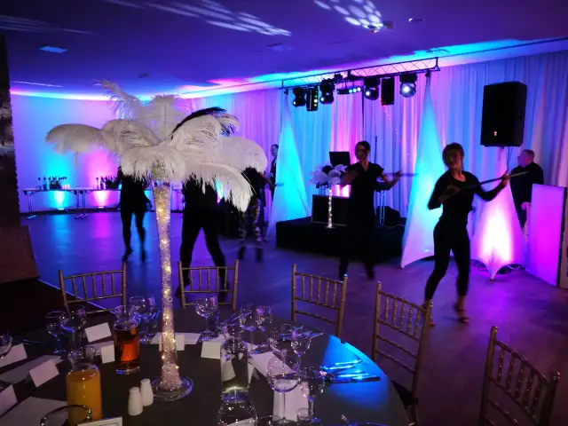 Impreza w stylu lat 20: tancerze, dekoracje z piór, stoły z kieliszkami i butelkami, kolorowe światła.