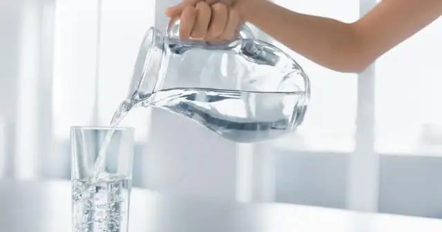 Dłoń przelewa czystą wodę alkaliczną z dzbanka do szklanki, tworząc bąbelki.