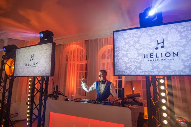 DJ w białej koszuli i granatowej kamizelce gra na weselu, na ekranach logo Helion Music Group.