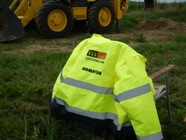 Żółta kamizelka z logo Komatsu i napisem "Szkolenia MM" leży na krześle. To przygotowanie do egzaminu na ładowarkę, gdzie pytania i odpowiedzi są kluczowe.