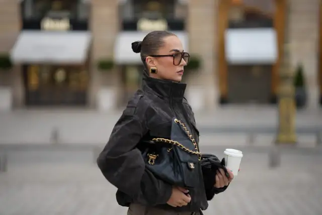 Elegancka kobieta w okularach i ciemnej kurtce puchowej, z torebką Chanel i kawą na wynos. Stylowe kurtki puchowe to podstawa jesiennych stylizacji.