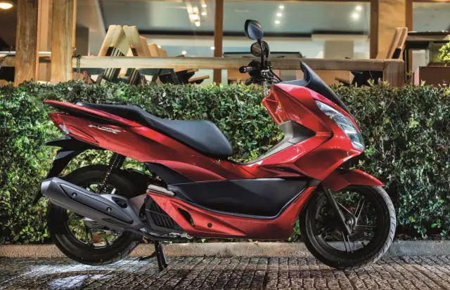 Czerwony skuter Honda PCX, jeden z popularnych **marek skuterów**, zaparkowany przed zielonym żywopłotem.