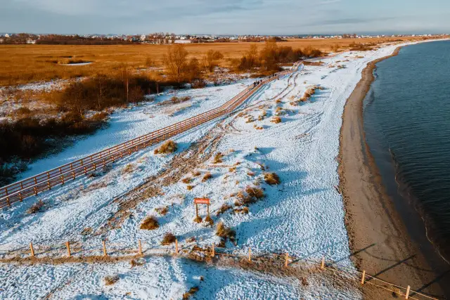 Zimowy krajobraz na rezerwacie przyrody Mechelińskie Łąki. Śnieg pokrywa plażę i wydmy, a drewniana kładka prowadzi wzdłuż morza.