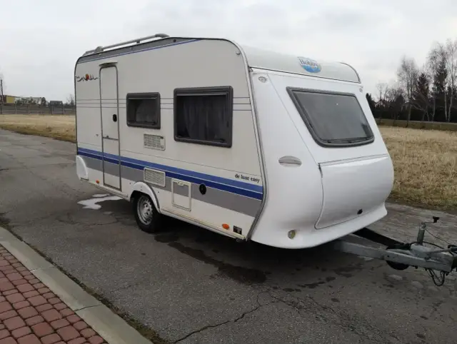 Biała przyczepa kempingowa Hobby 400 de luxe easy z niebieskimi pasami, gotowa na wakacyjne podróże.