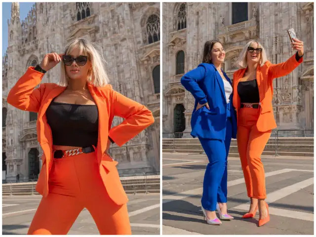 Dwie kobiety w stylizacjach z pomarańczowymi spodniami i marynarkami pozują na tle katedry. Jedna robi selfie, druga poprawia okulary.