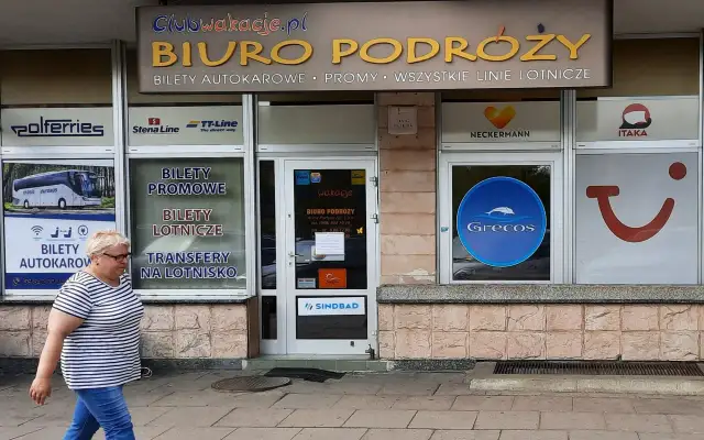 Biuro podróży z ofertą Grecos. Kobieta w paski idzie chodnikiem przed witryną z logo Grecos.