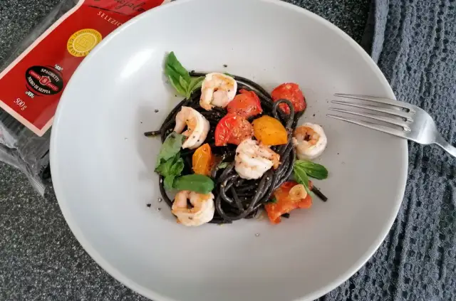 Czarny makaron z krewetkami, pomidorkami i bazylią na talerzu. Obok widoczna paczka spaghetti nero di seppia.