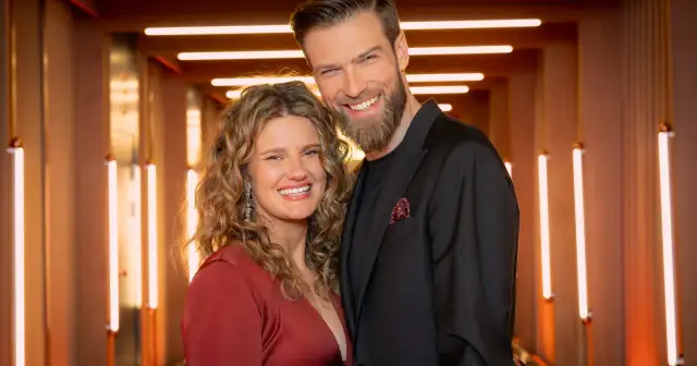 Uśmiechnięta para, jak z Love is Blind Polska, pozuje na tle rozświetlonego korytarza.
