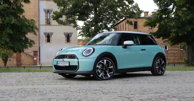 Turkusowy Mini Cooper stoi na brukowanej drodze przed ceglanym budynkiem.