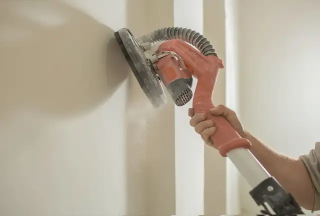 Ręka trzyma żyrafę do gipsu, która szlifuje ścianę. Pył unosi się w powietrzu.