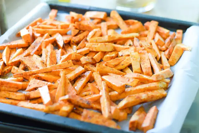 Frytki z batatów przyprawione i ułożone na blasze wyłożonej papierem do pieczenia.