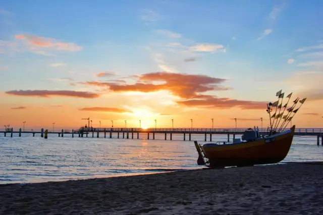 Łódź rybacka na plaży o zachodzie słońca, w tle molo i morze.