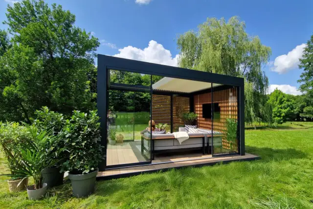Nowoczesny pawilon ogrodowy z przeszklonymi ścianami i wygodną sofą, idealny na relaks. Pokazuje, jak zrobić taras na trawie, tworząc przytulną przestrzeń.