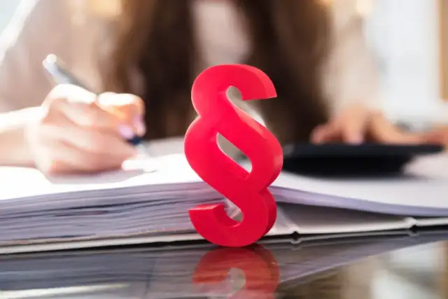 Czerwony paragraf symbolizuje prawne aspekty, gdy firma ogłasza upadłość, co z pracownikami? Tło pokazuje ręce pracujące przy dokumentach i kalkulatorze.