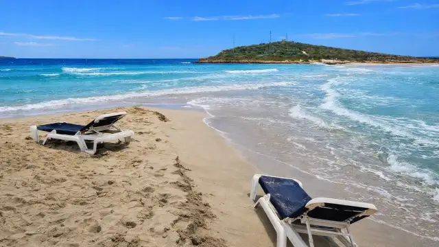 Plaża na Cyprze zimą. Dwa puste leżaki na piasku, turkusowe morze i zielone wzgórze w oddali.