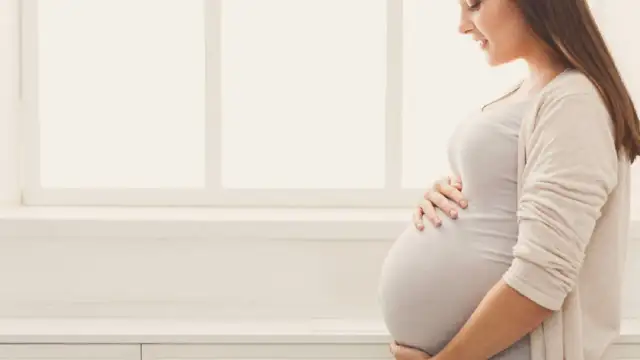 Uśmiechnięta ciężarna kobieta trzymająca się za brzuch na tle okna.