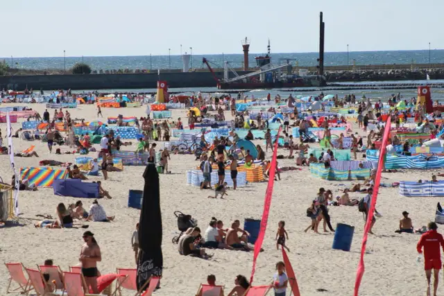 Tłum ludzi na plaży w Łebie. W tle widać morze i port. Tyle mieszkańców ma Łeba, że plaża jest tak pełna.