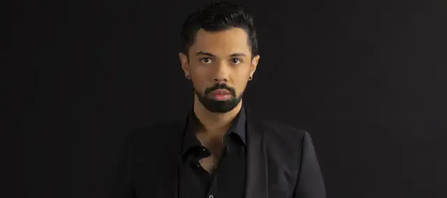 Mężczyzna w czarnym garniturze, z zarostem i kolczykiem w uchu. Jego spojrzenie sugeruje, że posiada najwyższy głos męski.