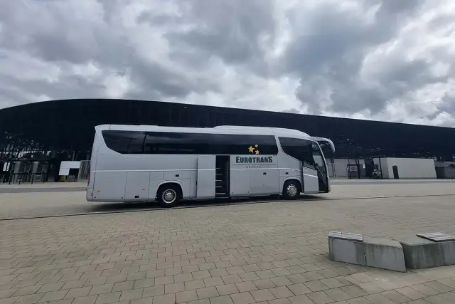 Biały autokar firmy Eurotrans z otwartymi drzwiami stoi na placu przed nowoczesnym budynkiem.