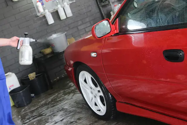 Ręka trzyma opryskiwacz, rozpylając pianę na czerwony samochód.
