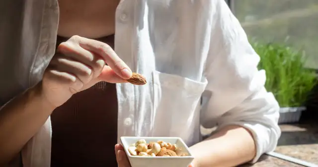 Osoba trzyma migdał, zastanawiając się, czy migdały szkodzą. W miseczce obok są inne orzechy.